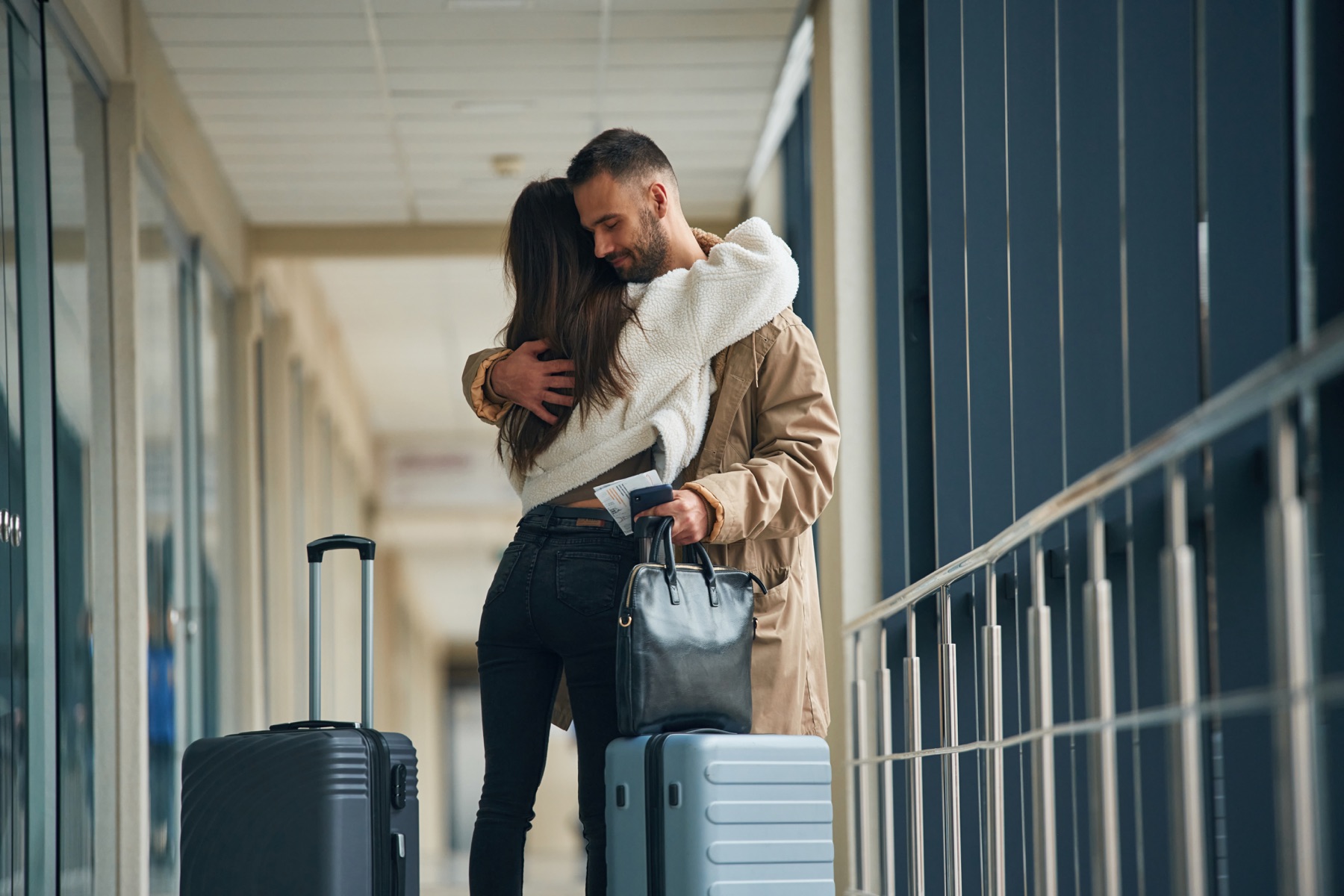 A couple hugging goodbye at the airport, suitcases beside them